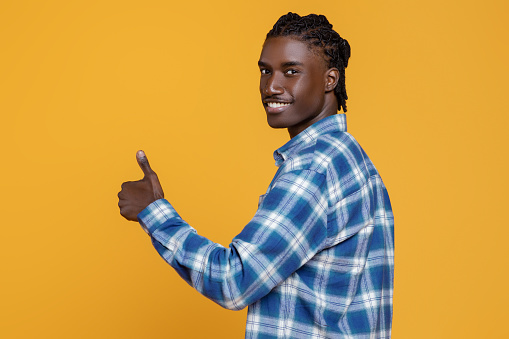 Smiling Young Black Guy Showing Thumb Up At Camera, Positive Millennial African American Man Standing Half-Turned On Yellow Studio Background, Gesturing Sign Of Approval, Recommending Something