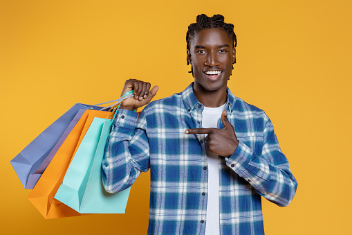 Cheerful Young Black Guy Pointing At Bright Paper Shopping Bags In His Hand, Happy Handsome African American Man Recommending Sales Offer, Standing Isolated On Yellow Studio Background