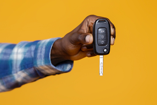 Closeup of black man's hand presenting set of car keys with remote, unrecognizable african american male suggesting ownership and mobility, standing isolated on yellow studio background, cropped
