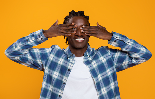 Happy young black man with braids hairstyle covering his eyes with hands, smiling millennial african american guy dressed in plaid shirt playing peekaboo game while standing on yellow background