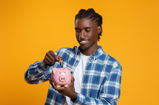 Smiling young black man with braids depositing coin into piggy bank, african american male showing financial responsibility, standing isolated on yellow studio background, copy space