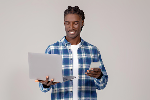 Focused young black man using laptop and holding smartphone, african american guy engaged in multitasking, wearing a casual plaid shirt, representing connectivity, standing on grey background
