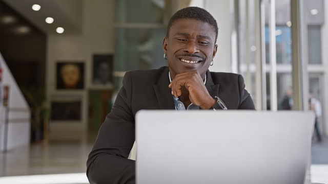 Smiling african man in suit using laptop in modern office interior, portrays professional work environment.