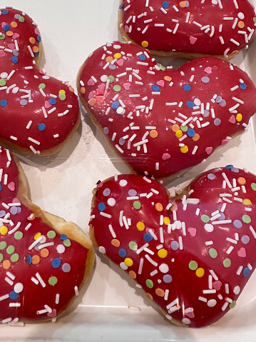 Stock photo showing close-up, elevated view of rows of red fondant iced heart doughnuts on white background.  The iced fried dough desserts are decorated with multicoloured hundred and thousand sugar sprinkles.