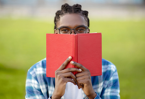Bookworm. Funny African American Student Guy Covering Face With Book, Peeking At Camera Wearing Eyeglasses, Preparing For Lectures Outdoors. Literature, Knowledge And Education