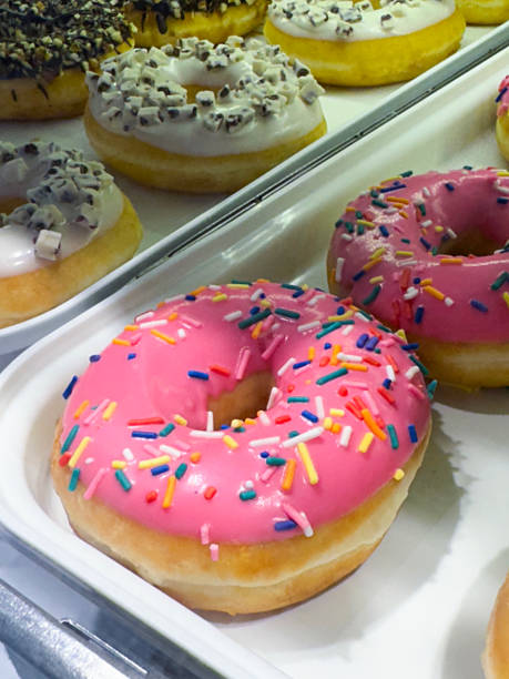istock Full frame image of plastic, white bakery trays displayed behind glass panel, rows of white and pink fondant iced and chocolate glazed ring doughnuts for sale, decorated with chocolate and multicoloured sugar sprinkles, elevated view, focus on foreground 2060249331