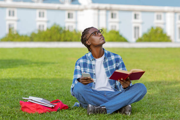 istock Thoughtful black guy reading book in park and using smartphone 2060249956