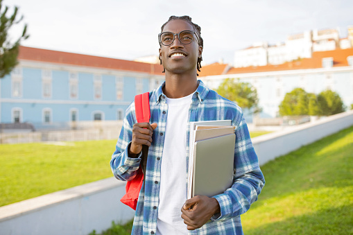 African American guy student in eyeglasses poses with laptop, backpack and his workbooks, standing near university building outdoors, smiling to camera with confidence. Joy of graduation