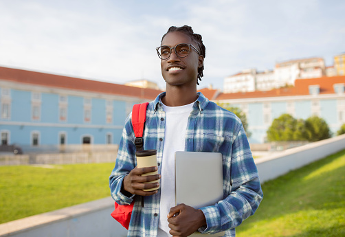 Happy African American college student guy stands holding laptop and coffee cup, blending leisure with learning outside at university park. E-learning and modern digital approach to education