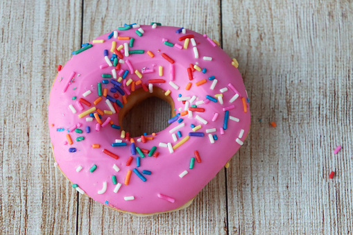 Stock photo showing elevated view of individual pink fondant iced ring doughnut on wood grain background.  The iced fried dough dessert is decorated with multicoloured hundred and thousand sugar sprinkles.