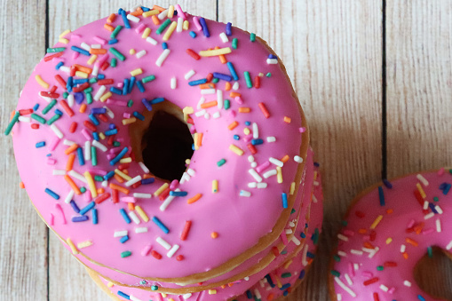 Stock photo showing close-up, elevated view of stack of pink fondant iced ring doughnuts, on wood grain surface, decorated with multicoloured hundred and thousand sugar sprinkles.