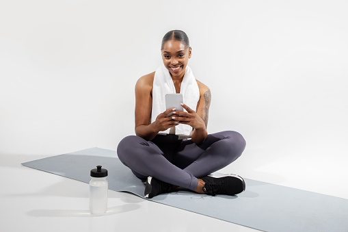 Black fitness woman browsing online workout on smartphone, taking break from training on mat, sitting with bottle of water and cellphone over white studio background. Sport applications