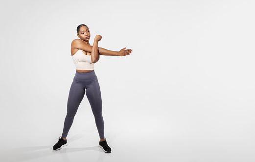 Full length shot of young black fitness woman stretching arms before training, warming up muscles having workout, standing over white studio background. Empty space for text