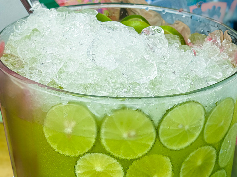 Stock photo showing close-up view of a glass jug containing lemon and lime juice with lime citrus fruit slices, ice cubes and crushed ice slushy covered in condensation showing the cold temperature of the beverage.