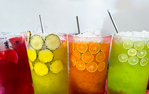 Stock photo showing close-up view of a row of four glasses containing watermelon, pineapple, orange, lemon and lime juices with watermelon, pineapple, orange and lime tropical and citrus fruit slices, ice cubes and crushed ice slushies covered in condensation showing the cold temperature of the beverage.