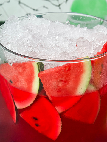 Stock photo showing close-up view of a glass containing watermelon juice with watermelon tropical fruit slices, ice cubes and crushed ice slushy covered in condensation showing the cold temperature of the beverage.
