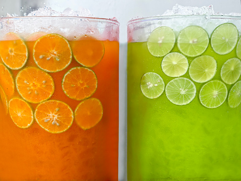 Stock photo showing close-up view of a glass jugs containing lemon and lime juice with lime citrus fruit slices and orange juice with orange slices, ice cubes and crushed ice slushy covered in condensation showing the cold temperature of the beverage.