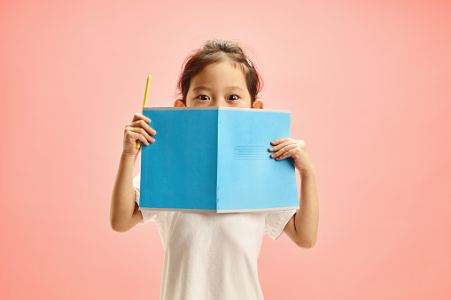 Isolated image of Asian ethnicity kid opening eyes to widen, hiding face behind an open notebook, looking at the camera in fright, was faced with a difficult or unsolvable task, confused standing over soft pink background with an empty copy space for elementary grade educational promotional