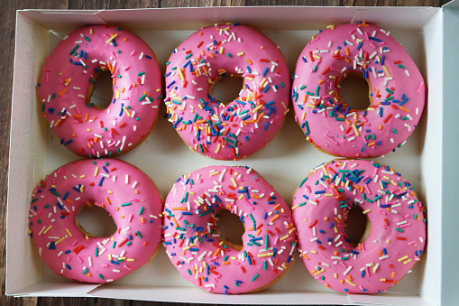 Stock photo showing close-up, elevated view of rows of pink fondant iced ring doughnuts in cardboard box.  The iced fried dough desserts are decorated with multicoloured hundred and thousand sugar sprinkles.