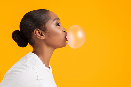 Side view portrait of black young woman blowing bubble, looking aside at copy space on yellow studio background. Cool millennial African American lady chewing gum having sweet tooth