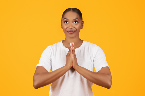 Faith. Portrait Of Smiling African American Lady Praying With Clasped Hands, Religious Young Black Woman Standing And Praying With Hope Over Yellow Studio Background