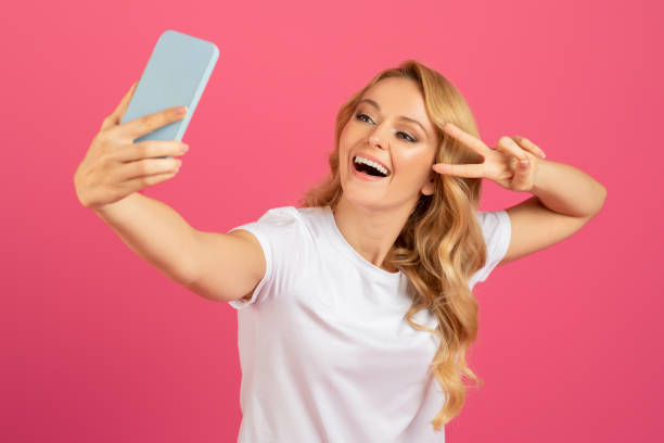 istock blonde woman making selfie on smartphone gesturing victory sign, studio 2060287786