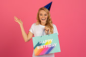 blonde woman in party hat holding Happy Birthday bag, studio