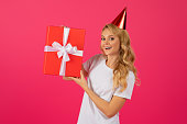 Happy blonde woman wearing birthday hat holding gift, pink background