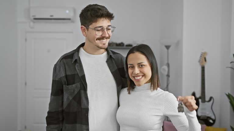 A happy couple, man and woman, posing in a modern living room, with the woman holding house keys.