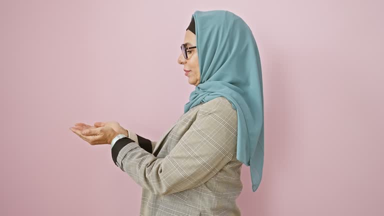 Excited middle-aged hispanic woman in hijab cheerfully pointing aside with open palm, presenting ad space over isolated pink wall, happily welcoming your business!