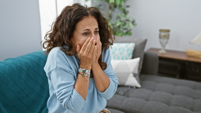 Stunned middle-age hispanic lady, sitting on her home sofa, covering her face in shock with her hand, peeking through her fingers, displaying a mix of fear and surprise.