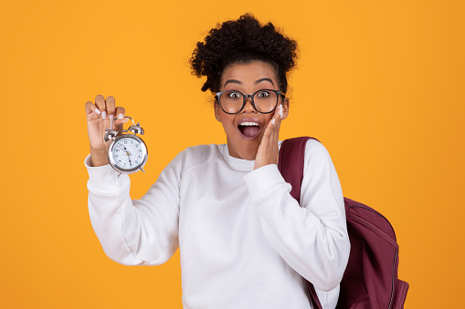 Shocked young black woman with backpack holding an alarm clock, emotional african american female student late to study, touching face with shock, standing isolated against vivid yellow background