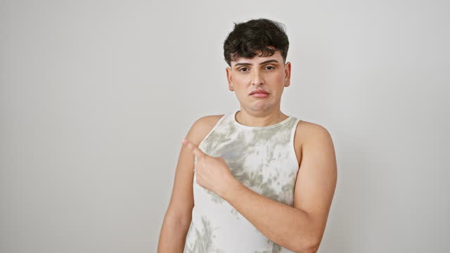 Worried young man in a sleeveless t-shirt, nervously pointing aside with forefinger on white isolated background, revealing shock and surprise.