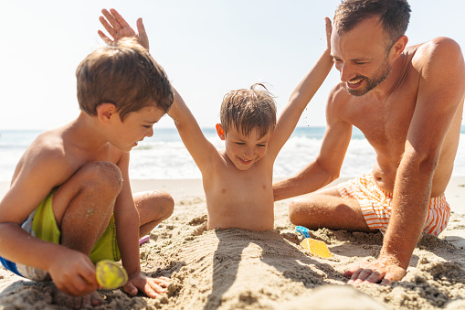 Photo of a single father with two young boys, spending quality time together on the beach. Boys are digging up the sand and playing with it.