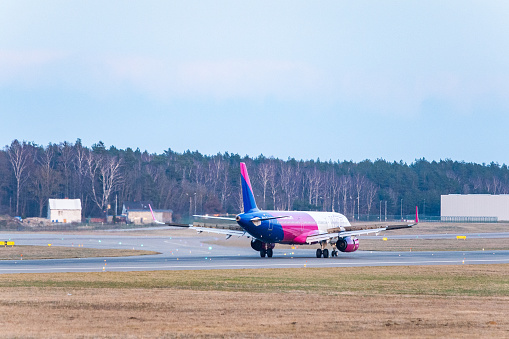 Gdansk - Lech Walesa Airport, a pink and blue airplane of Wizz Air (Airbus A320) is taxiing on the runway of an airport. The background is a clear blue sky. The foreground is a grassy area with some runway lights.