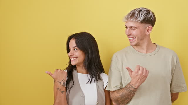 Joyful couple standing together, smiling, pointing to the side with a thumbs up gesture, expressing their optimistic and cheerful emotions over isolated yellow wall background.