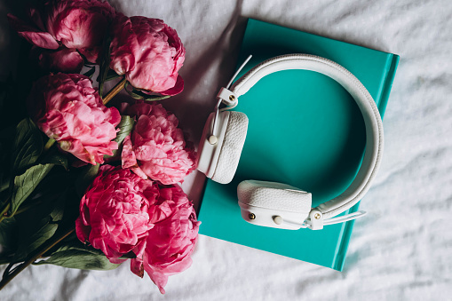 Book, white headphones and a bouquet of spring pink peony flowers on the bed, top view. Audiobook, reading and relaxation concept.