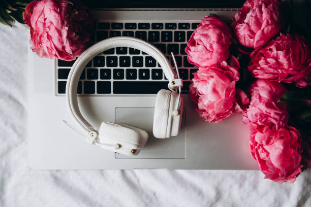 White headphones, laptop and pink peonies on white sheets in bed, top view.