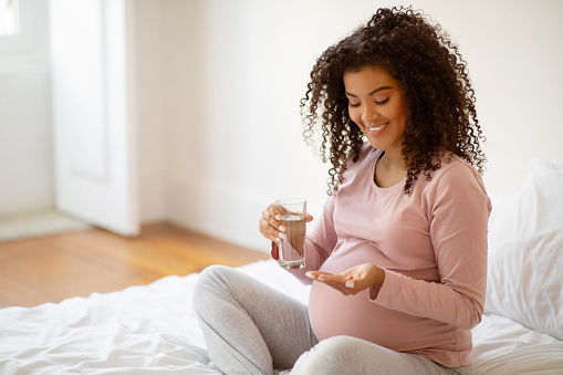 Prenatal Vitamins. Beautiful Smiling Black Pregnant Woman Holding Pills And Glass Of Water, African American Expectant Mother Taking Supplements For Healthy Pregnancy, Sitting On Bed At Home