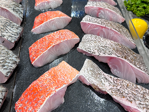 Stock photo showing close-up, elevated view of a retail seafood display with sea bass, red grouper and red snapper steaks on chilled metal tray.