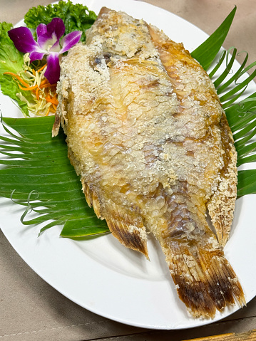 Stock photo showing close-up, elevated view of a white, oval plate containing a whole, cooked grouper fish which was baked in salt applied to the skin.