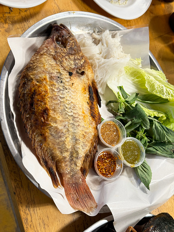 Stock photo showing close-up, elevated view of a metal, circular tray containing a whole, cooked grouper fish which was baked in salt applied to the skin.