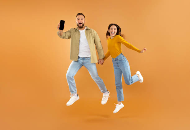 istock Couple Jumping And Showing Smartphone With Empty Screen, Orange Wall 2060390224