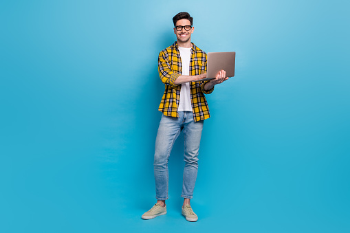 Full size body photo of young guy wear yellow shirt with jeans and going to work using macbook laptop isolated on blue color background