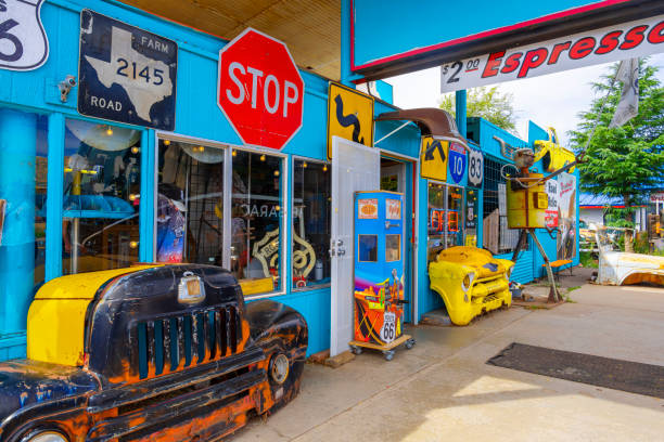Lamar, Colorado, USA - May 31, 2024: Vintage vehicles, including a yellow bus, populate a rustic outdoor space under large trees. Various old car parts and equipment scatter the area, creating a nostalgic atmosphere. Dusty ground and weathered automobiles contribute to the unique ambiance of this vintage car dealership or junkyard