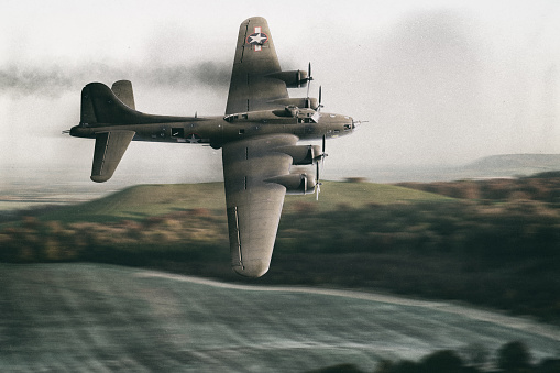 With smoke streaming out of one engine, a (scale model) battle-damaged B-17 Flying Fortress flies at a low altitude over a green landscape. The B-17 was an American four-engined heavy bomber aircraft developed in the 1930s for the United States Army Air Corps and made by Boeing.