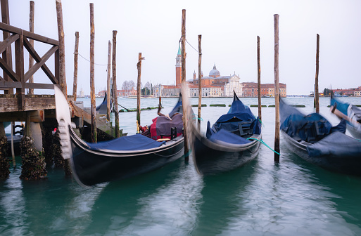 Gondolas tied up on the side of a misty Grand Canal in Venice. Across the water we see the iconic shape of the Church of San Giorgio Maggiore