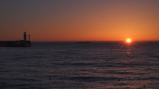 Sunrise at sea. Ships and a lighthouse can be seen on the horizon.