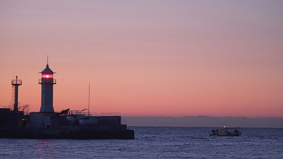 Fishing boats sail near the lighthouse. Sunrise at sea.