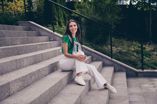 Photo of cheerful glad lovely woman dressed stylish clothes sitting on stairs in park spending summer days outdoors.